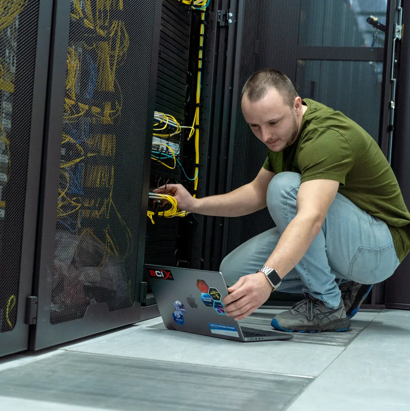 Mitarbeiter in grüner T-Shirt und Jeans hockt vor einem Laptop mit Aufklebern, konfiguriert Server in einem Rechenzentrum mit sichtbaren gelben Kabeln.